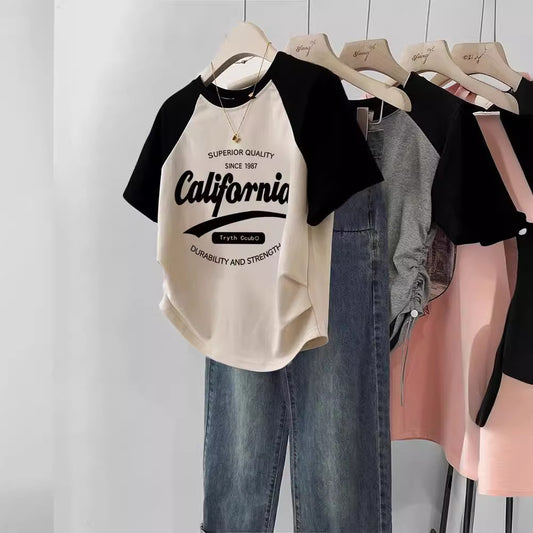 Beige and black baseball shirt on a hanger with jeans and pink skirt in the background.